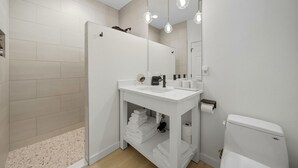 A modern bathroom with a white sink, mirror, and toilet. There are hanging lights above the mirror and a towel rack below the sink.