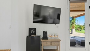 A modern living room with a flat-screen TV mounted on the wall, a black refrigerator, a wooden cabinet with drawers, and a view of an outdoor patio with seating.