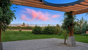 A covered outdoor area with a gravel floor, surrounded by greenery and a view of distant mountains.