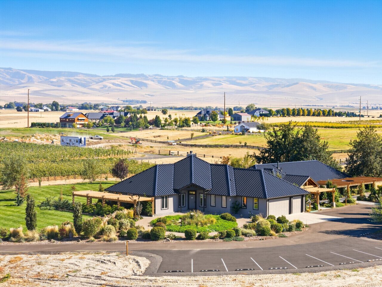 A modern building with a dark roof, surrounded by a parking lot and a vineyard.