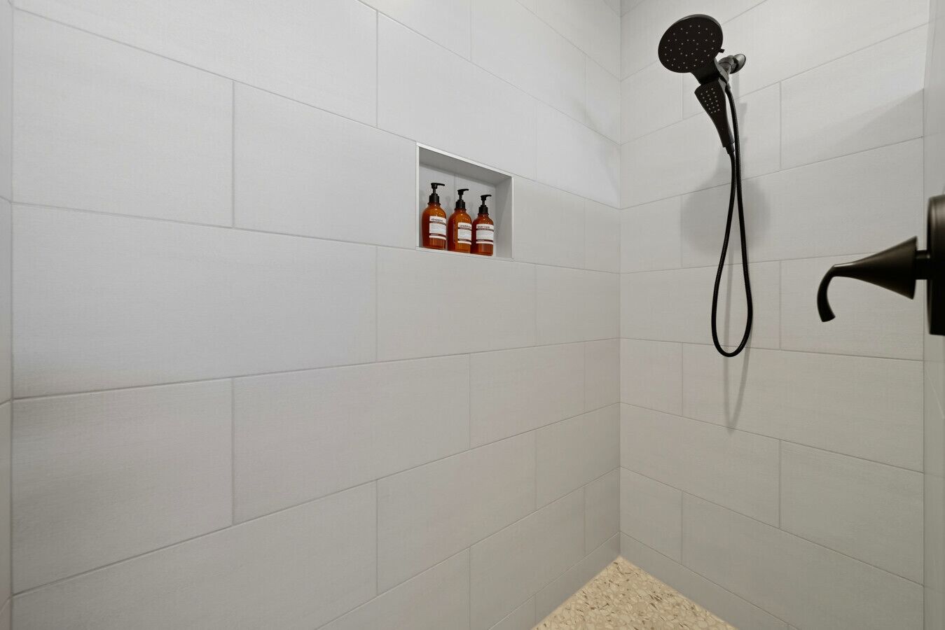 A shower area with white tiles, a handheld showerhead, and a shelf with three bottles.