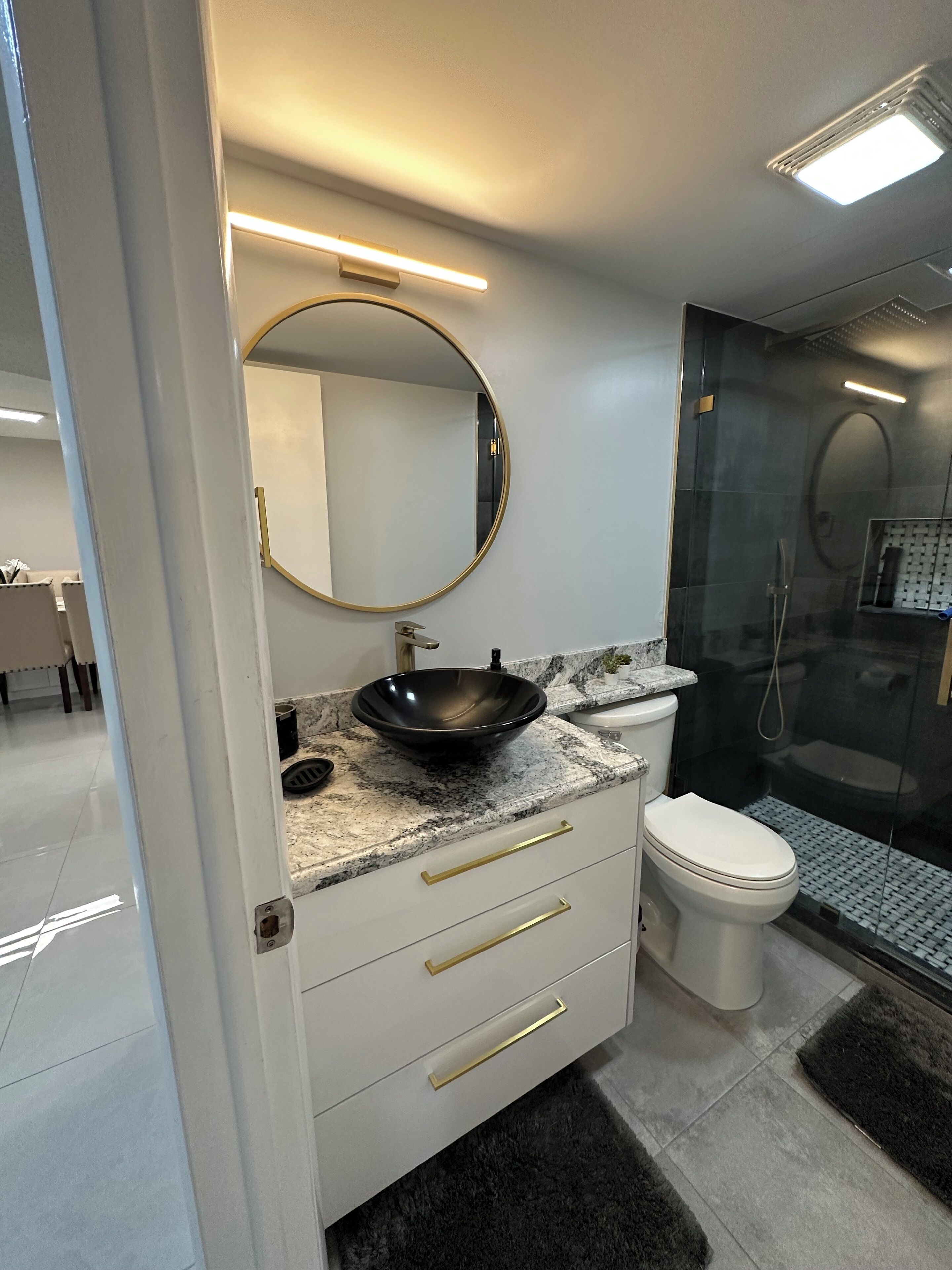 A modern bathroom with a round mirror, a black vessel sink, a white toilet, and a glass-enclosed shower.