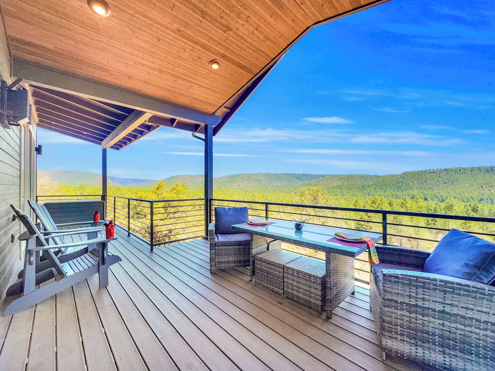 A covered outdoor deck with wicker furniture, a table, and chairs, overlooking a forested landscape.
