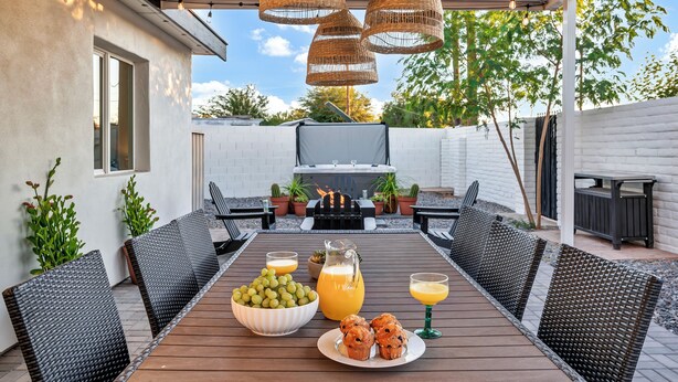 Eine Terrasse im Freien mit Holztisch, der zum Essen gedeckt ist, Korbstühlen, einem Grill und Topfpflanzen.