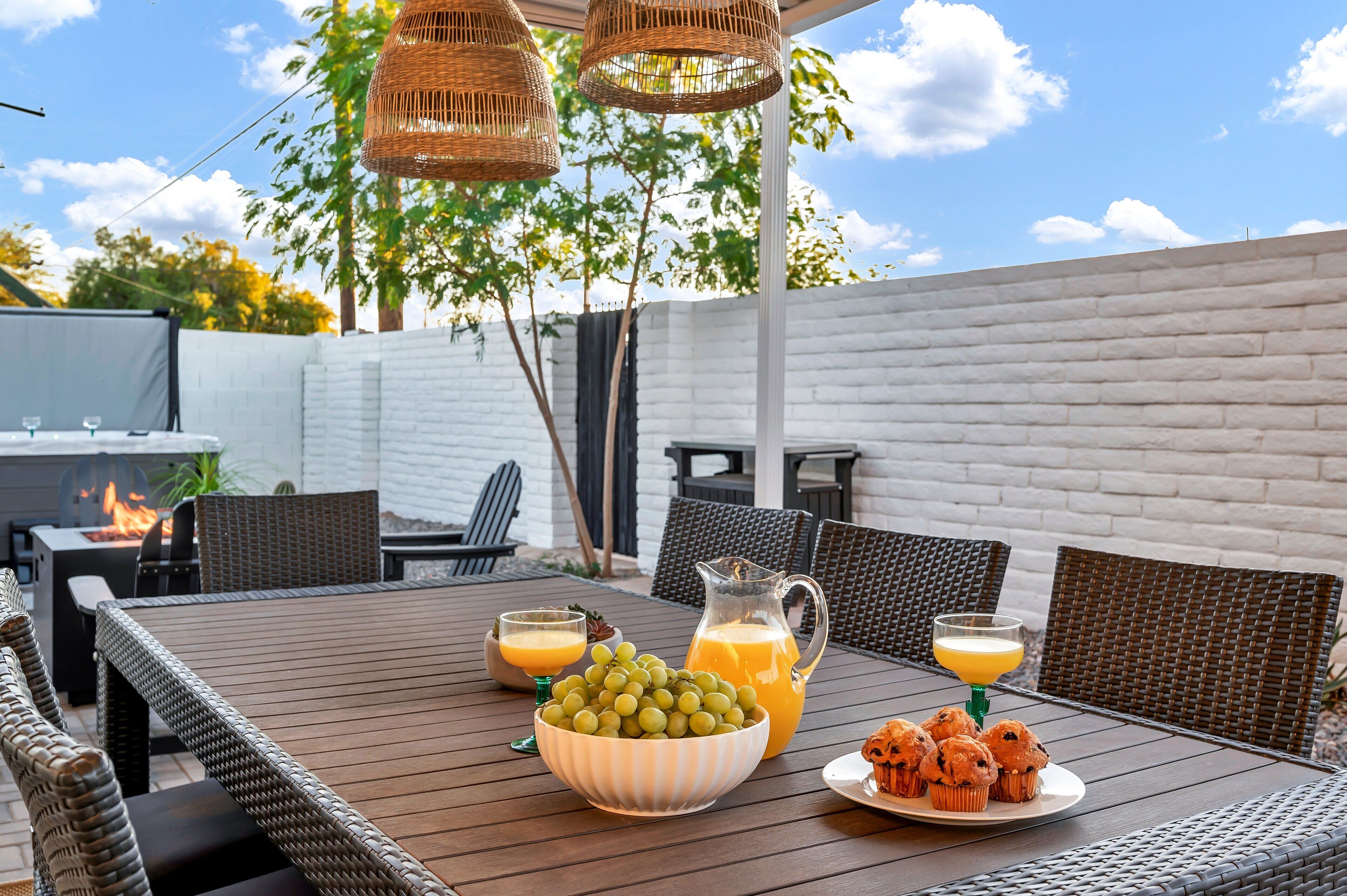 An outdoor patio with wicker furniture, a table with a bowl of grapes and muffins, and a pitcher of orange juice.