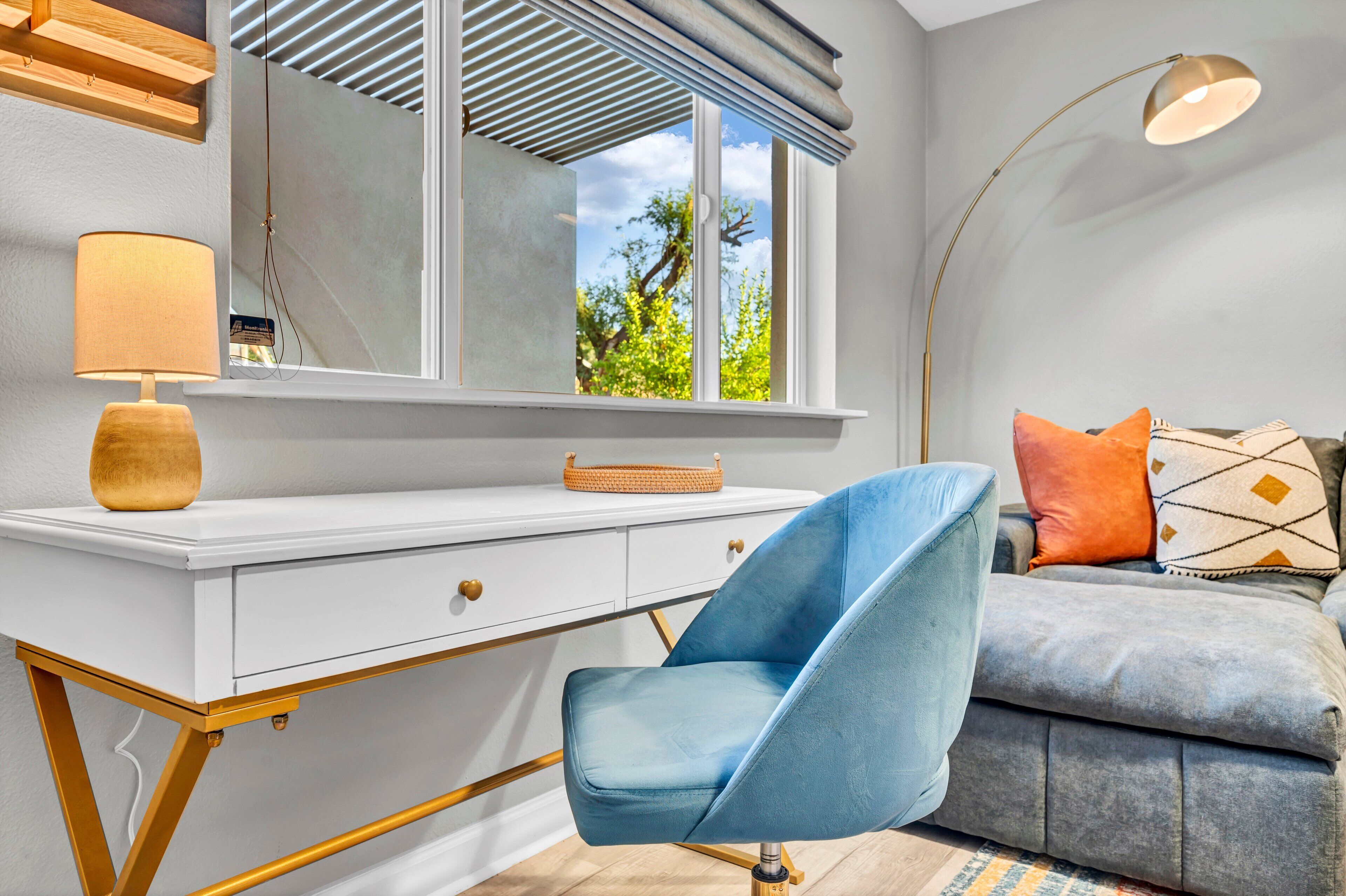 Een moderne kamer met een wit bureau, een blauwe stoel, een bank met kleurrijke kussens, een vloerlamp en een raam met jaloezieën.