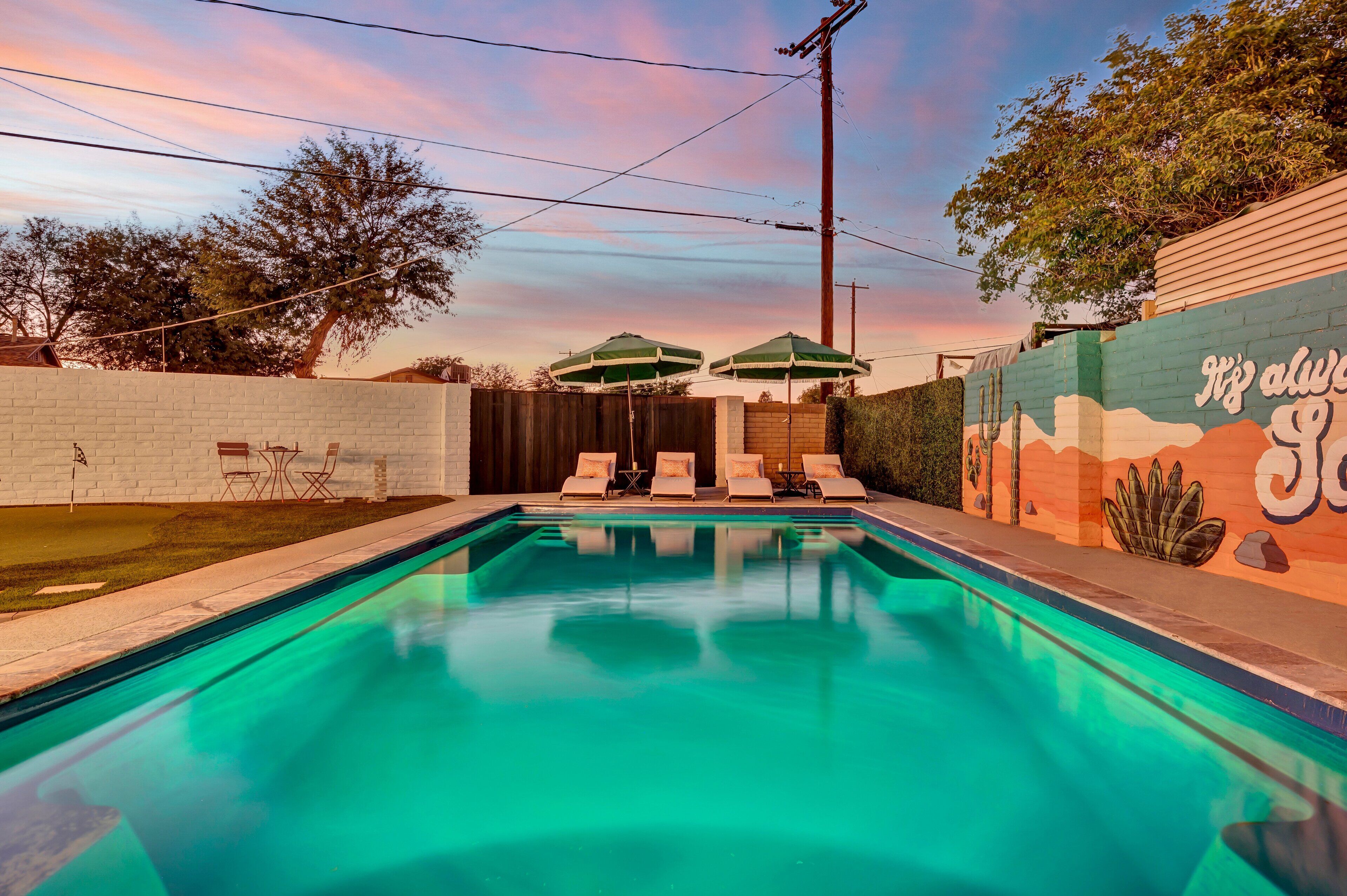 Una piscina con sillas de descanso y sombrillas, rodeada por un muro de ladrillo con un mural.