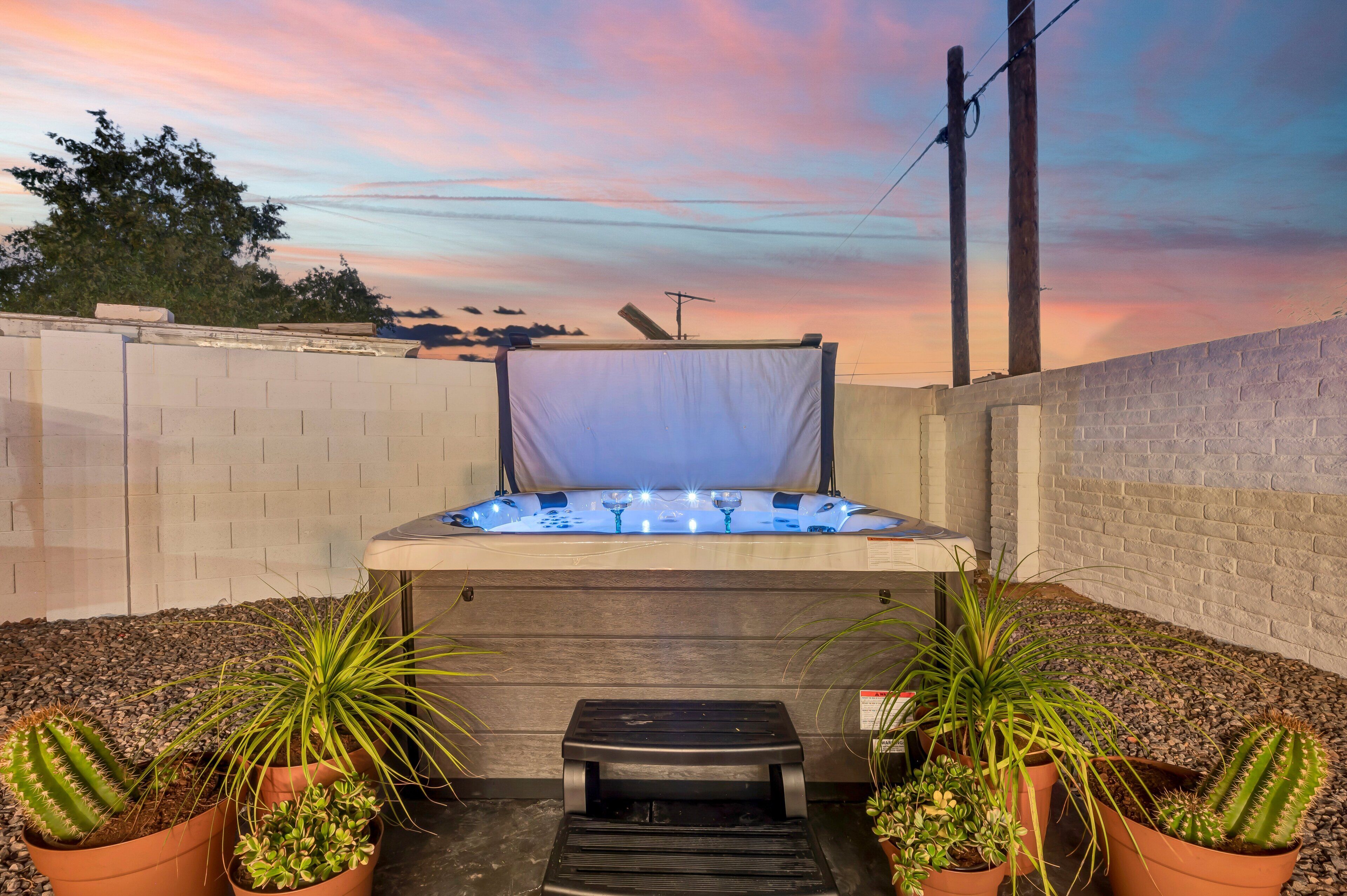A hot tub in a backyard with a brick wall, potted plants, and a sunset sky.