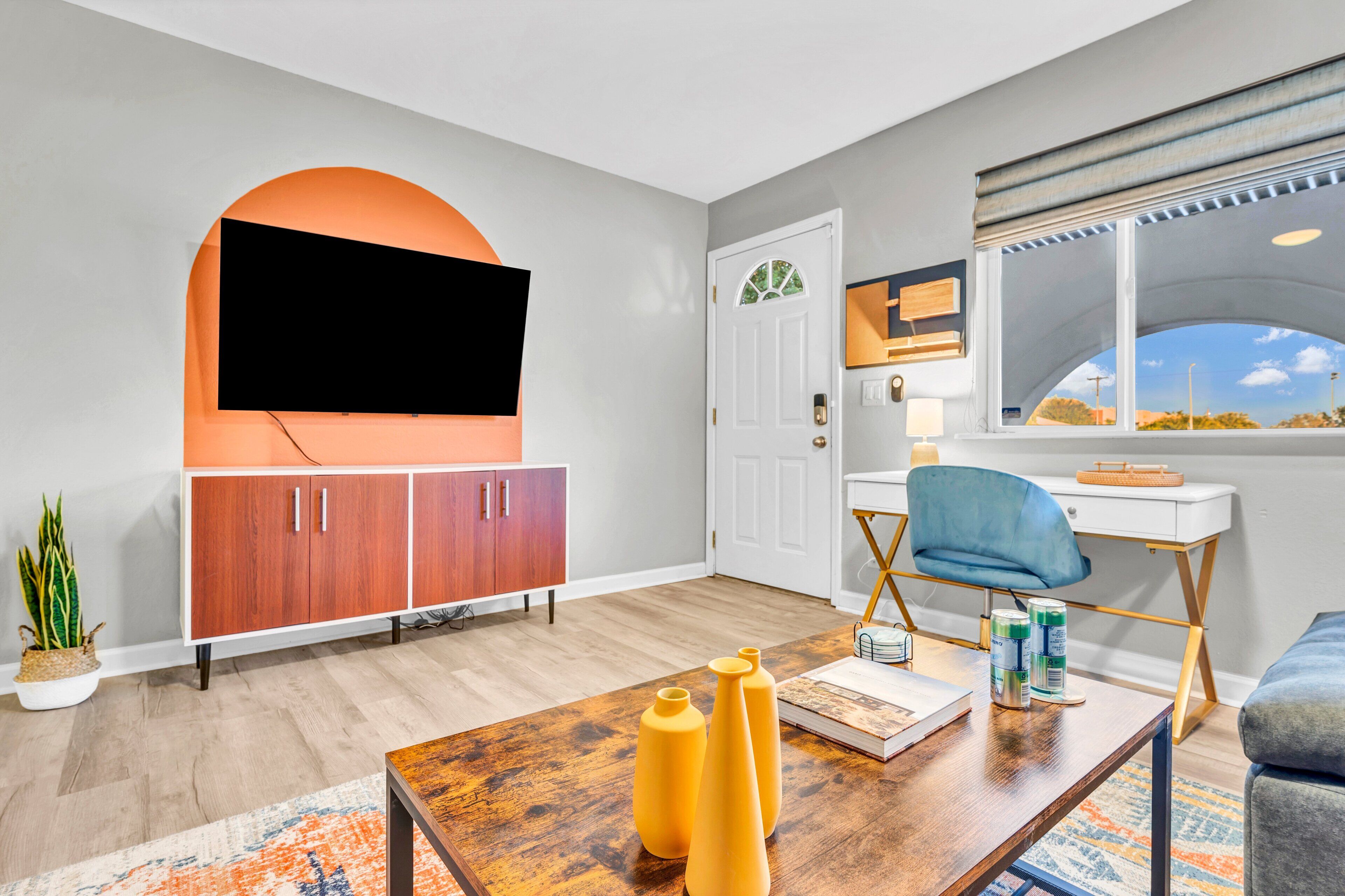 Una sala de estar moderna con un televisor de pantalla plana montado en la pared, una mesa de centro de madera con libros y objetos decorativos, un sillón azul y una ventana con vistas al exterior.