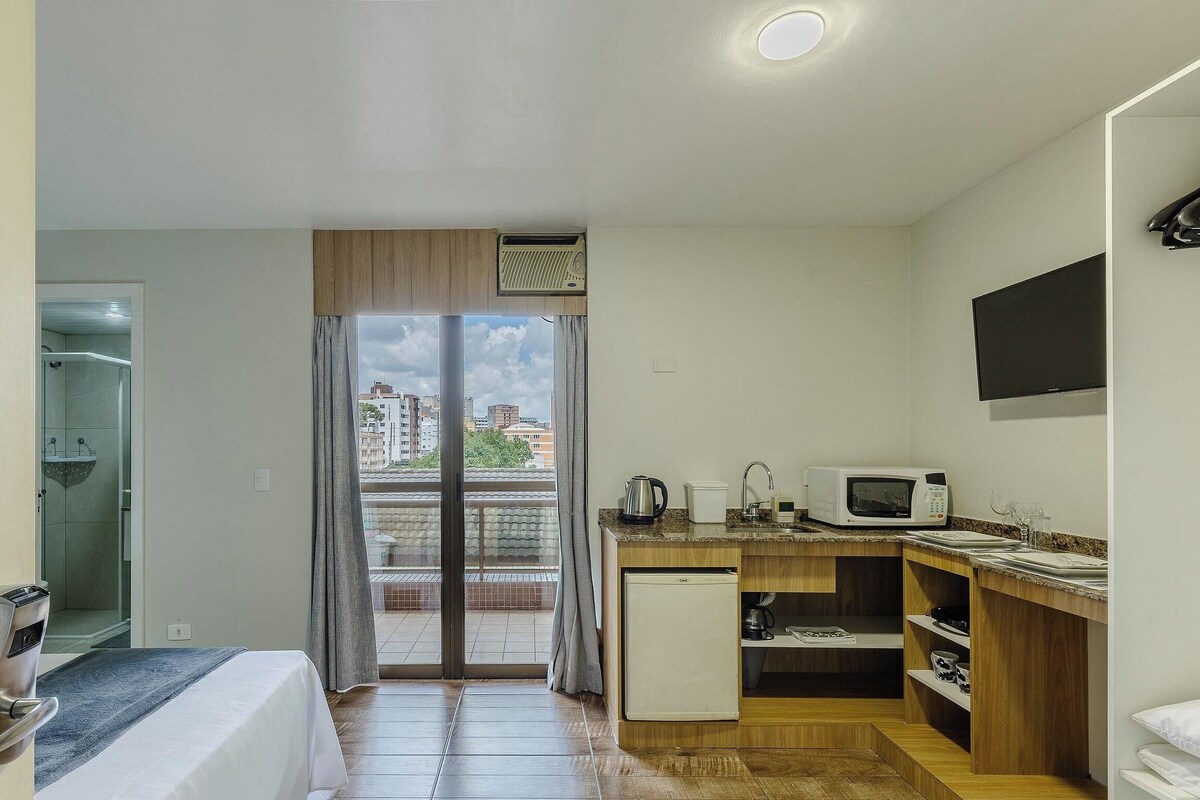 Camera d'albergo compatta con un letto, angolo cottura, piccolo frigorifero, forno a microonde e vista sulla città tramite una portafinestra.