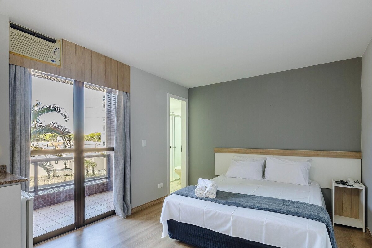 A bedroom with a bed, a bedside table with a phone, a sliding glass door leading to a balcony, and a view of buildings outside.