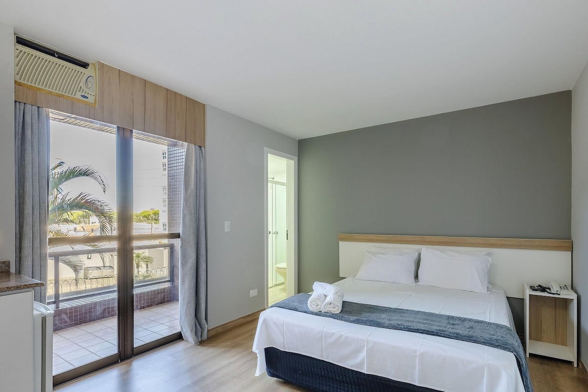 Een slaapkamer met een bed, een nachtkastje met een telefoon, een schuifglazen deur die leidt naar een balkon, en uitzicht op gebouwen.