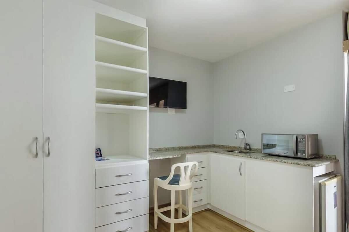 Een kleine keuken met witte kastjes, een magnetron, een kleine koelkast en een stoel.