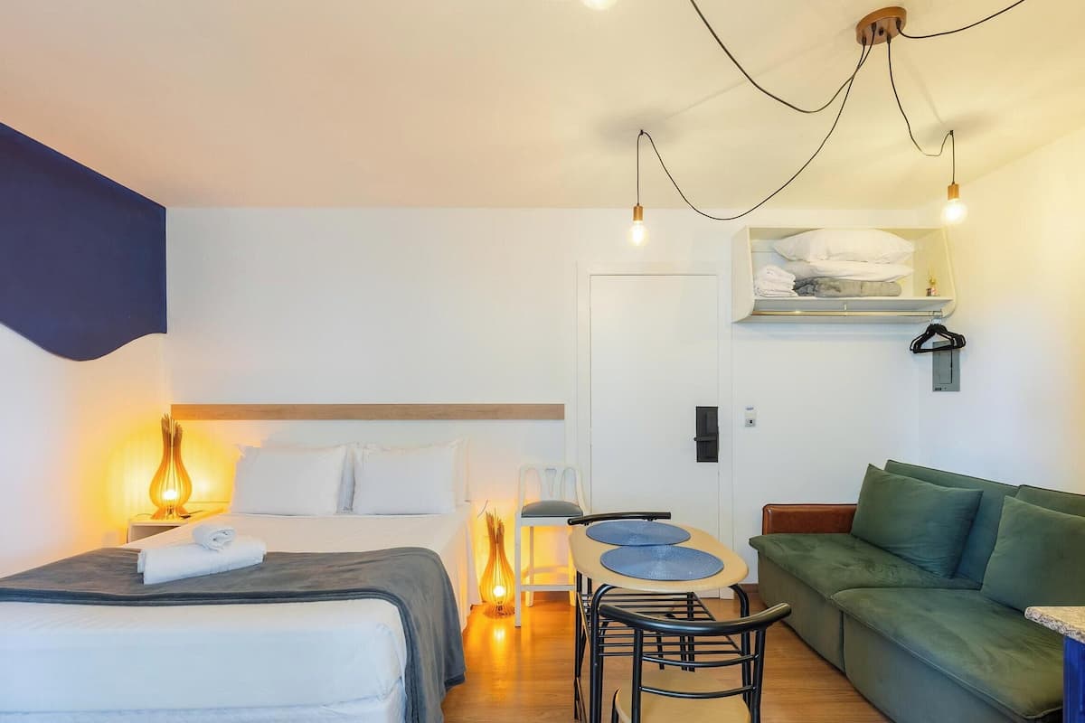 Een hotelkamer met een bed, een groene bank, een kleine eettafel en een stoel. Er zijn wandlampen en een plafondlamp.