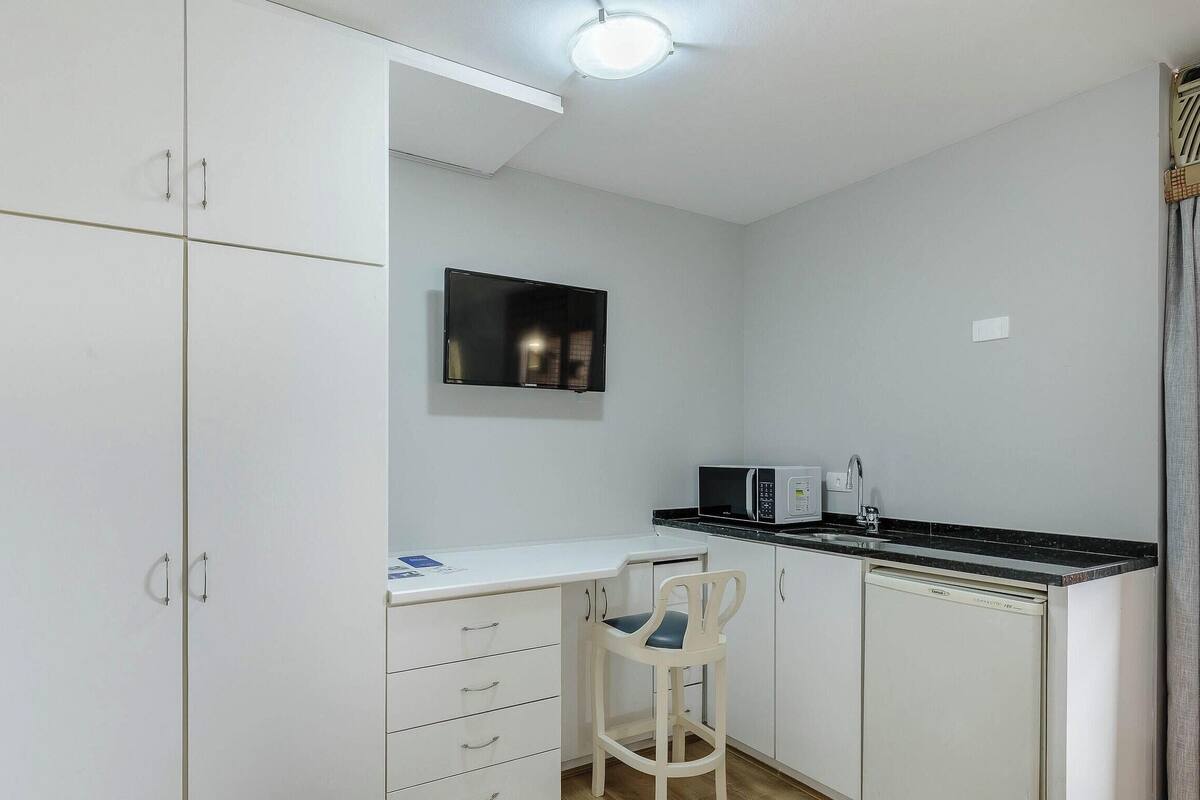 Een kleine keuken met witte kastjes, een magnetron, een spoelbak en een stoel.