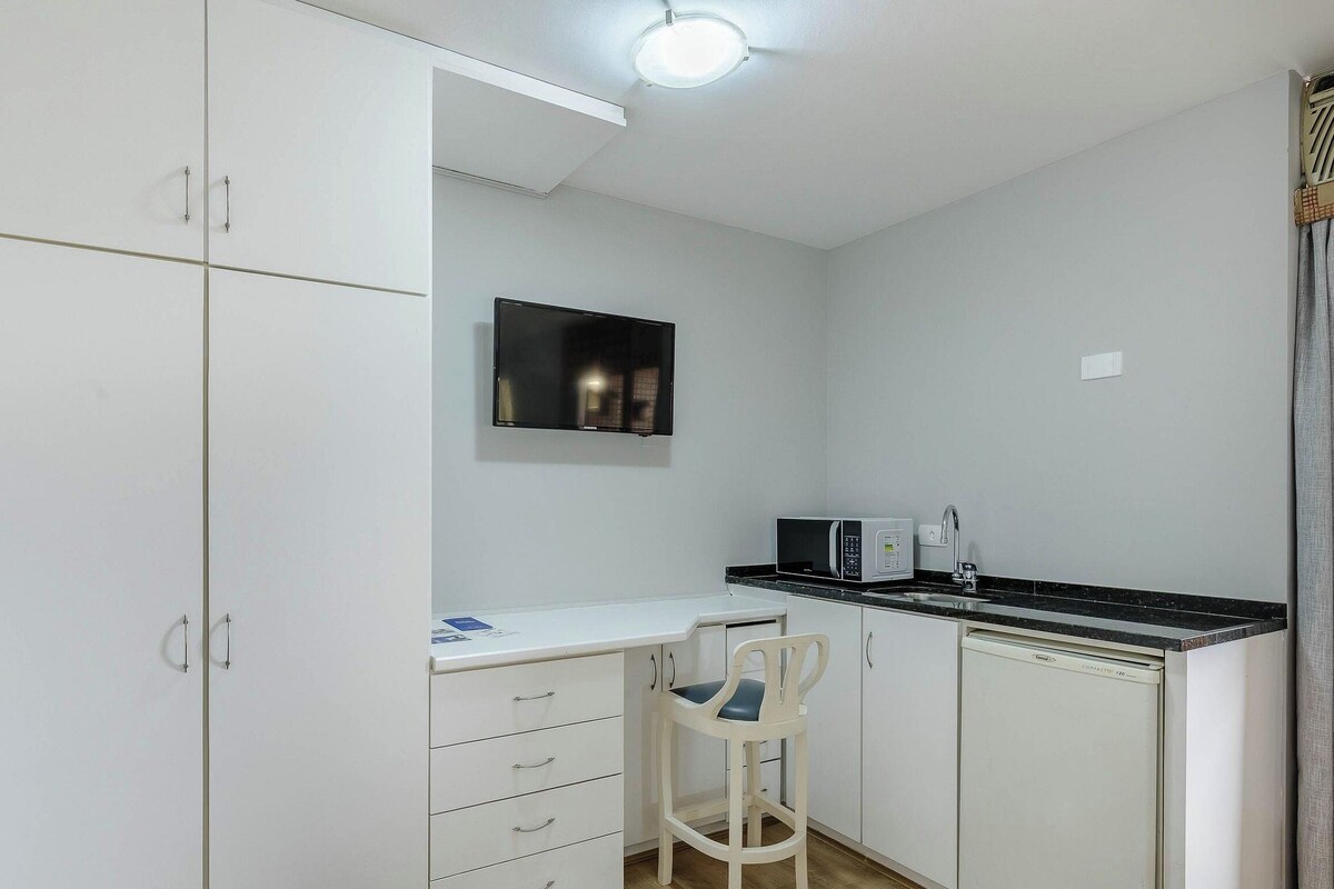 Een compacte keuken met witte kasten, een zwart aanrechtblad, een magnetron en een klein eethoekje met een stoel.