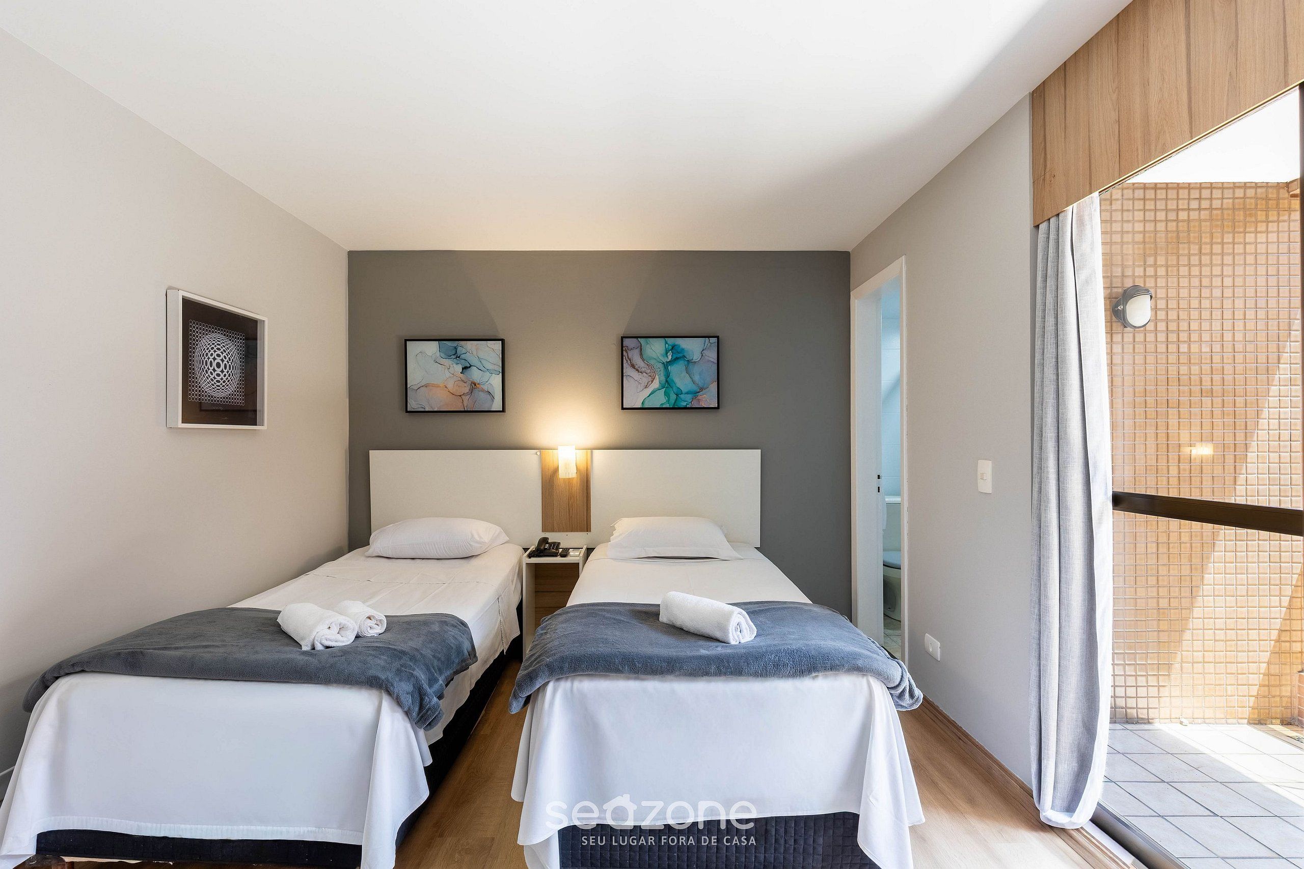 A hotel room with two beds, a nightstand, a wall-mounted lamp, and a large window with a view.