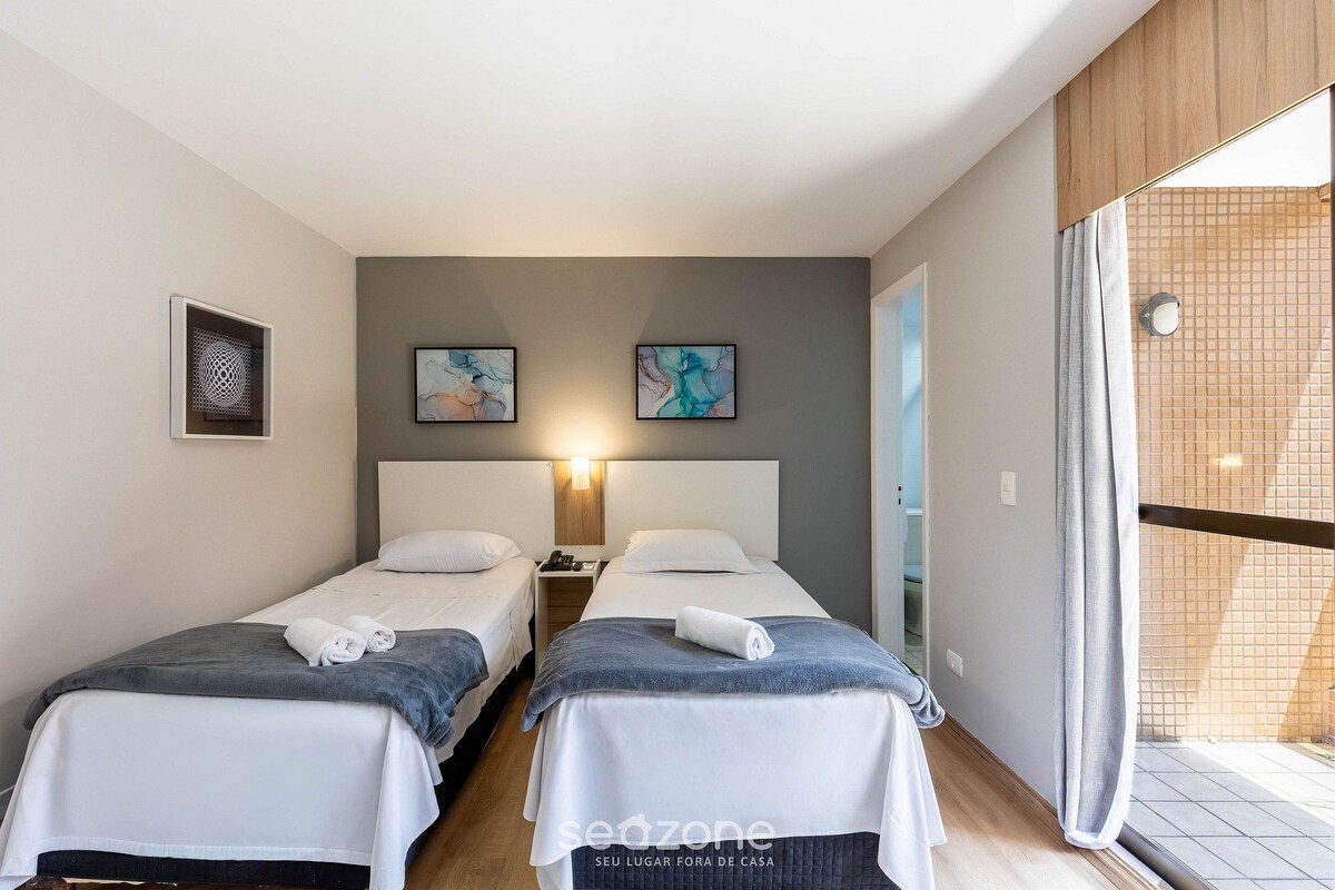 Camera d'albergo con due letti, un comodino, una lampada a parete e un'ampia finestra con vista.