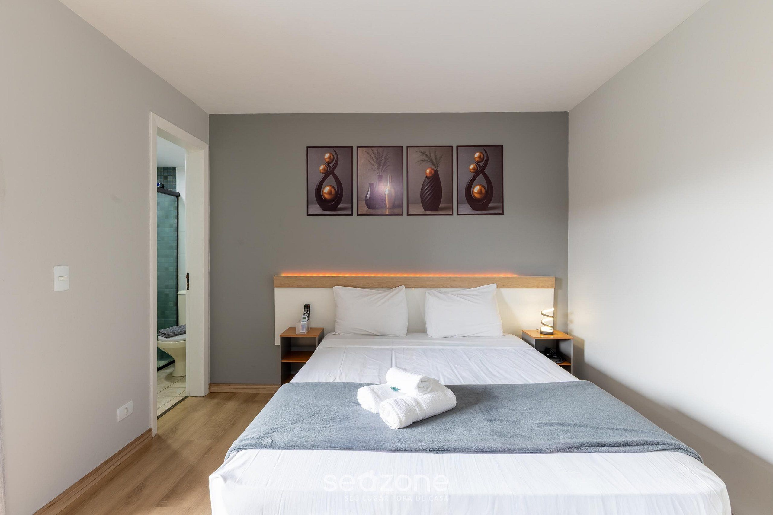 Une chambre d’hôtel avec un lit, une table de chevet, une œuvre d’art accrochée au mur et une salle de bain visible à travers une porte ouverte.