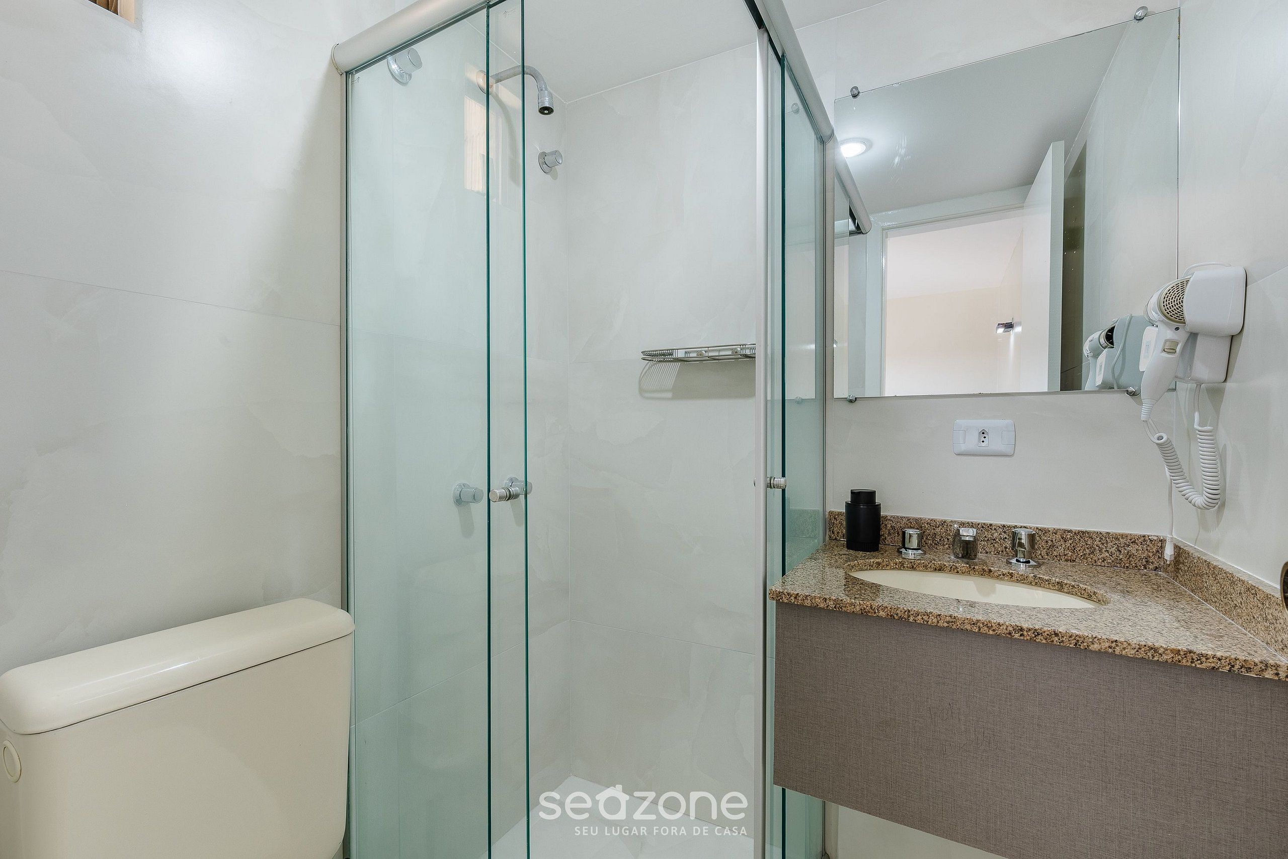 A bathroom with a glass shower enclosure, a white toilet, a sink with a granite countertop, and a hairdryer mounted on the wall.