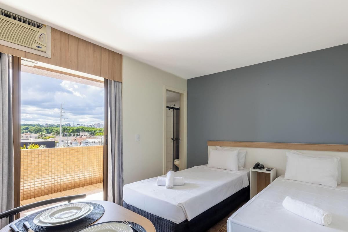 Een hotelkamer met twee bedden, een kleintje tafeltje met bord en vork, een telefoon op een nachtkastje en uitzicht op de stad door een groot raam.