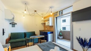 A compact hotel room with a green sofa, a small dining table, a kitchenette, a large window, and a potted plant. - Studios at Upper Residence - UPP (Curitiba)