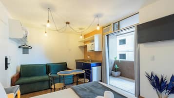 A compact living space with a green sofa, a small dining table, a kitchenette, and a sliding glass door leading to a balcony.