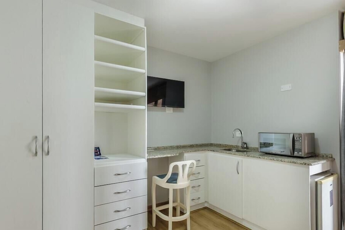 A compact kitchen with white cabinets, a microwave, a small refrigerator, and a chair.