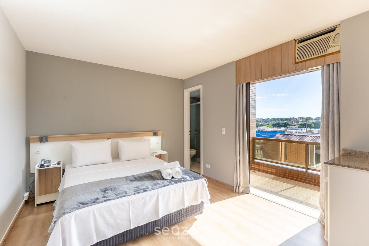 A bedroom with a bed, bedside tables, a bathroom door, and a balcony with a view.