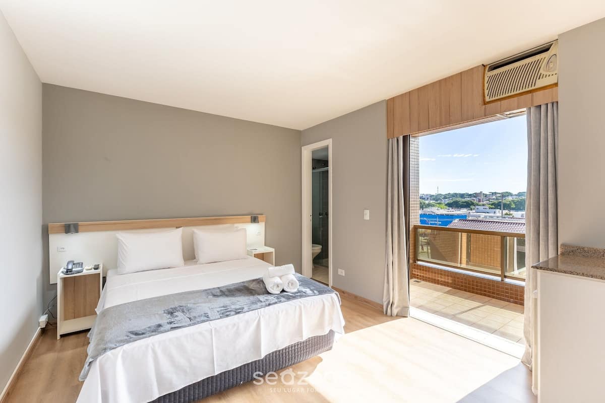 Een slaapkamer met een bed, nachtkastjes, een badkamers deur en een balkon met uitzicht.