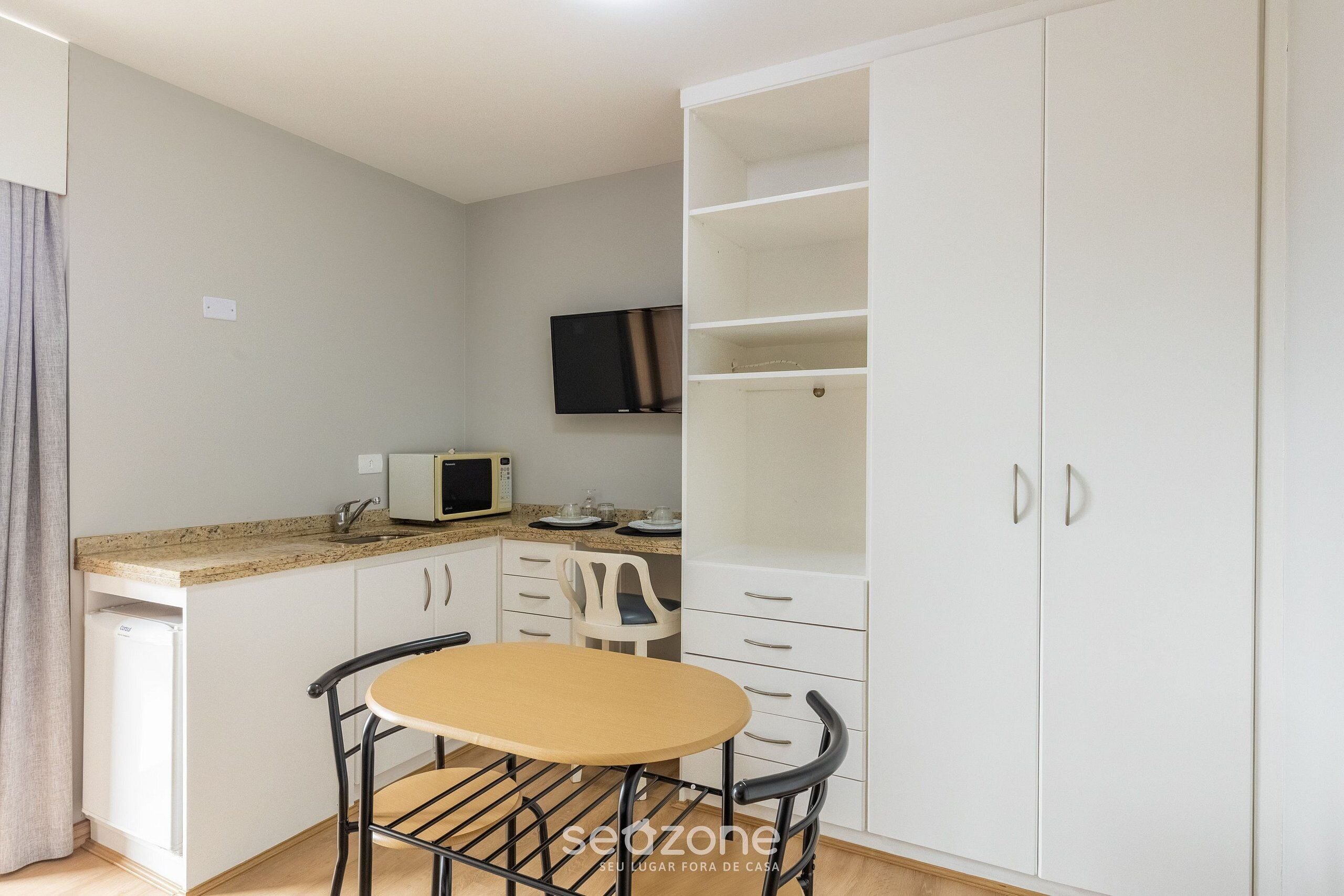 A compact kitchen with a microwave, white cabinets, a small table, and a TV mounted on the wall.