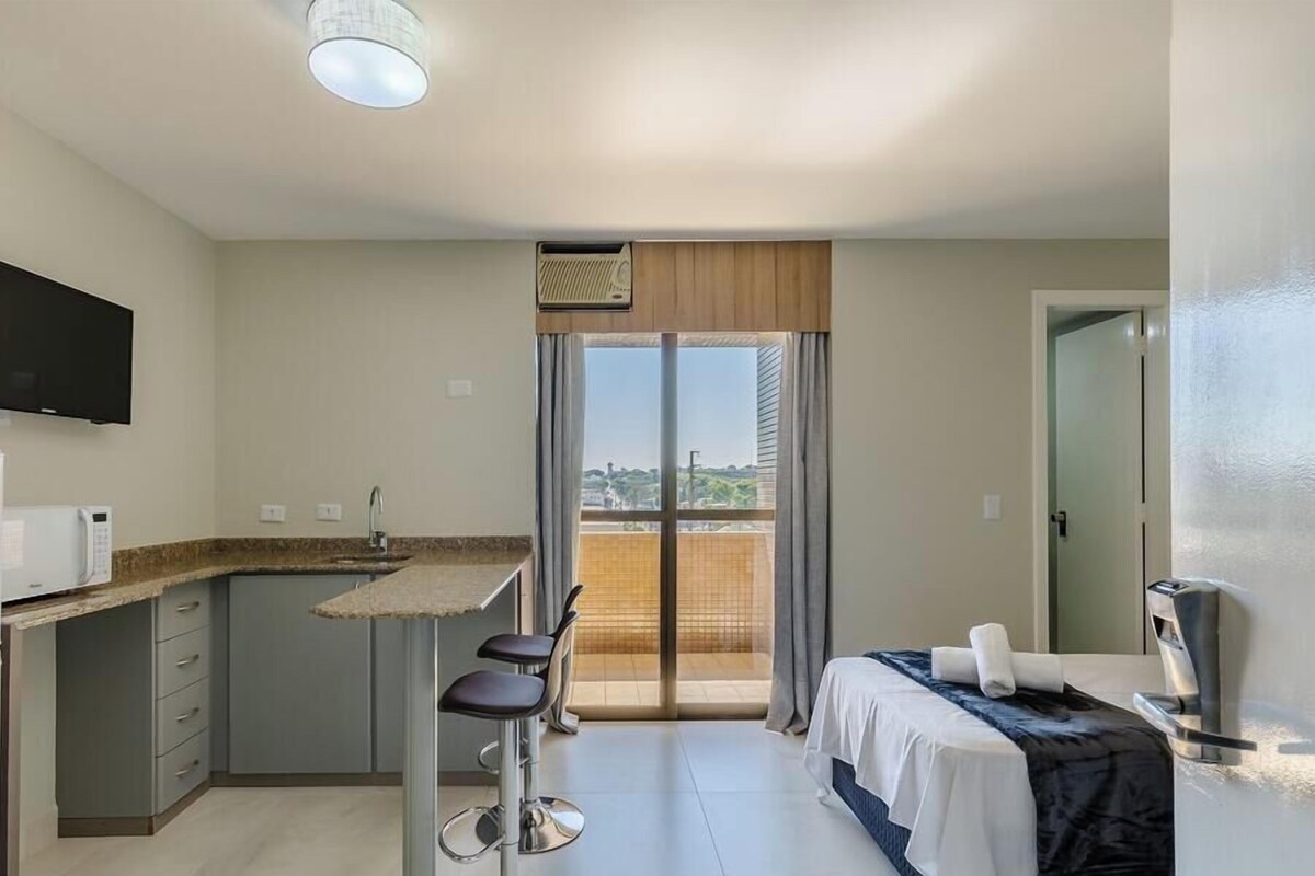 A compact hotel room with a kitchenette, a bed, a TV, and a balcony with a view.