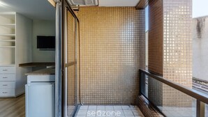 A modern interior with a tiled wall, a small refrigerator, a flat-screen TV, and a balcony with a railing. - Studios at Upper Residence - UPP (Curitiba)