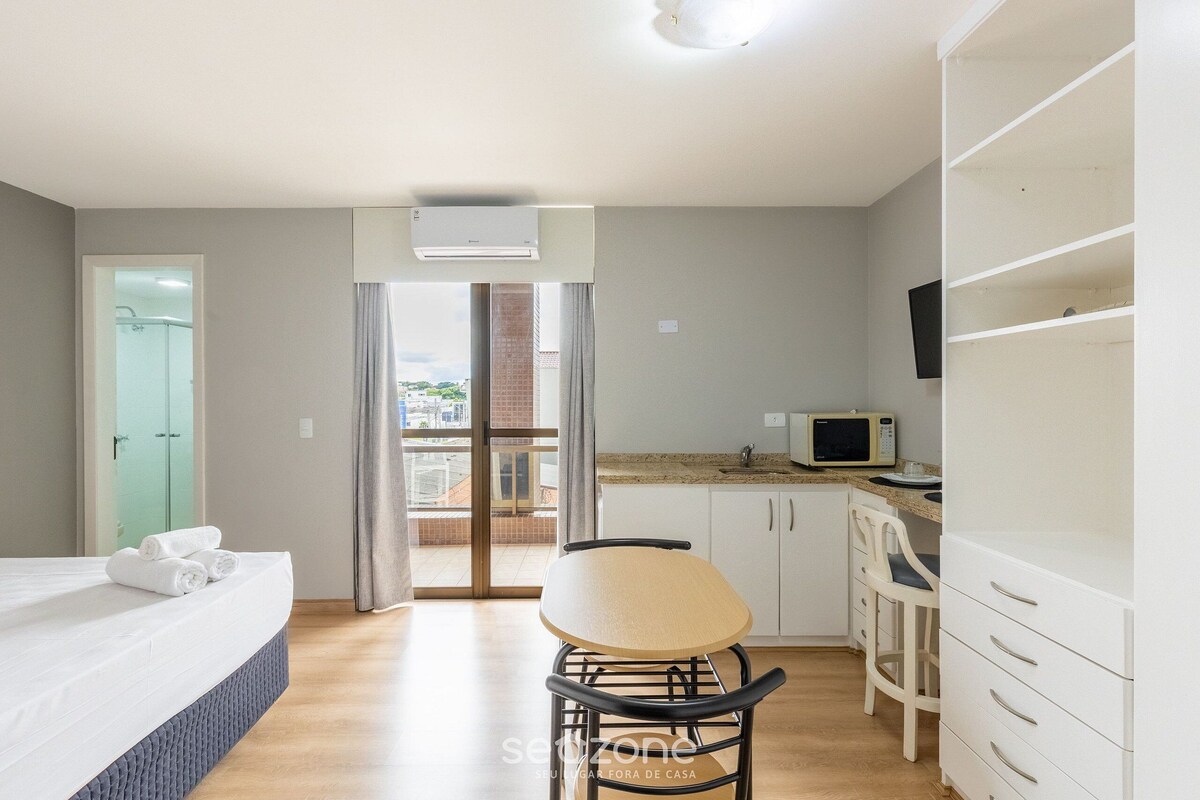 A room with a bed, a chair, a kitchen area, and a balcony with a view.