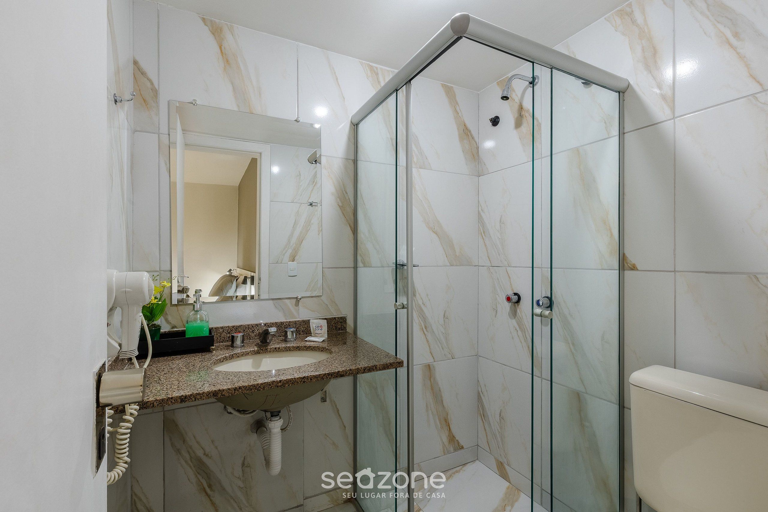 A bathroom with a glass shower enclosure, a marble-tiled wall, a sink with a granite countertop, and a toilet.