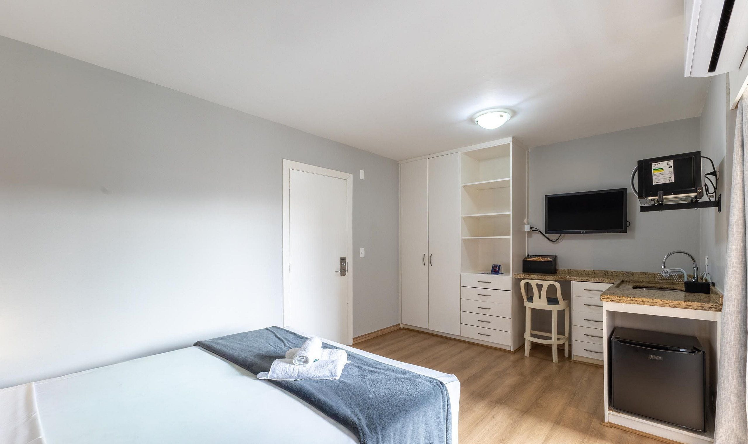 Un dormitorio con cama, escritorio con silla, televisor y una pequeña kitchenette.
