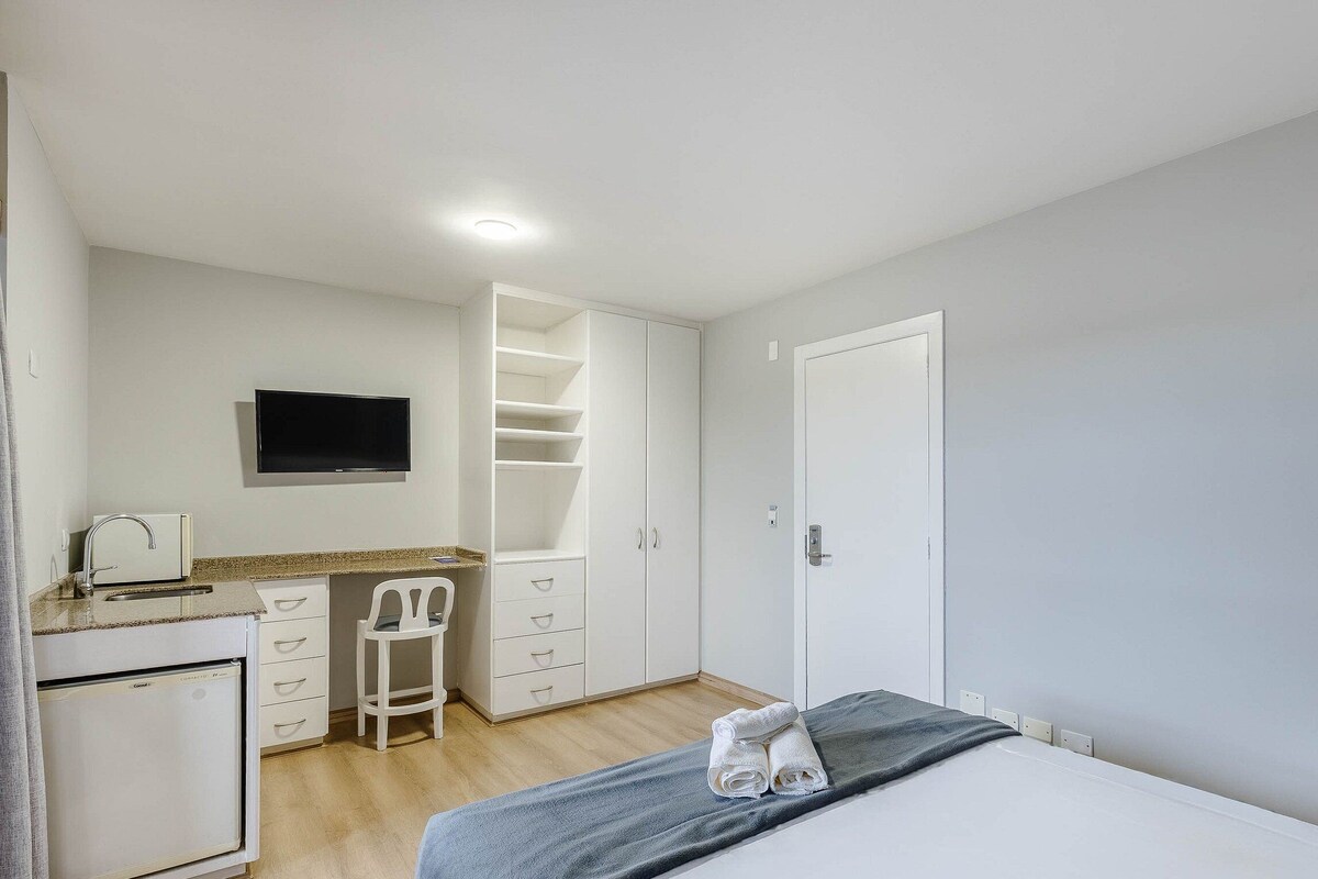 A bedroom with a bed, a desk with a chair, a TV mounted on the wall, and a wardrobe.
