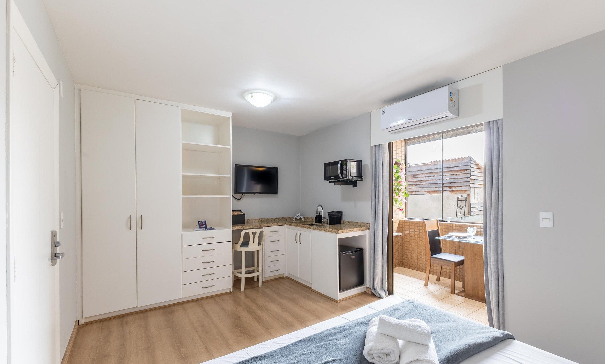 A compact room with a kitchenette, a bed with towels, a TV, and a view of the outside.