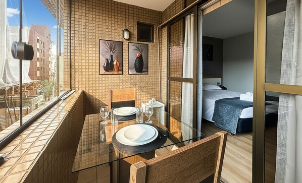 A balcony with a table set for two, a bed in the adjacent room, and a cityscape view.