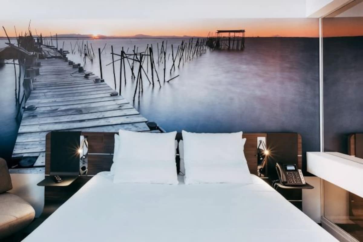 Una habitación de hotel con cama, mesitas de noche y vista a un muelle que se adentra en el agua.