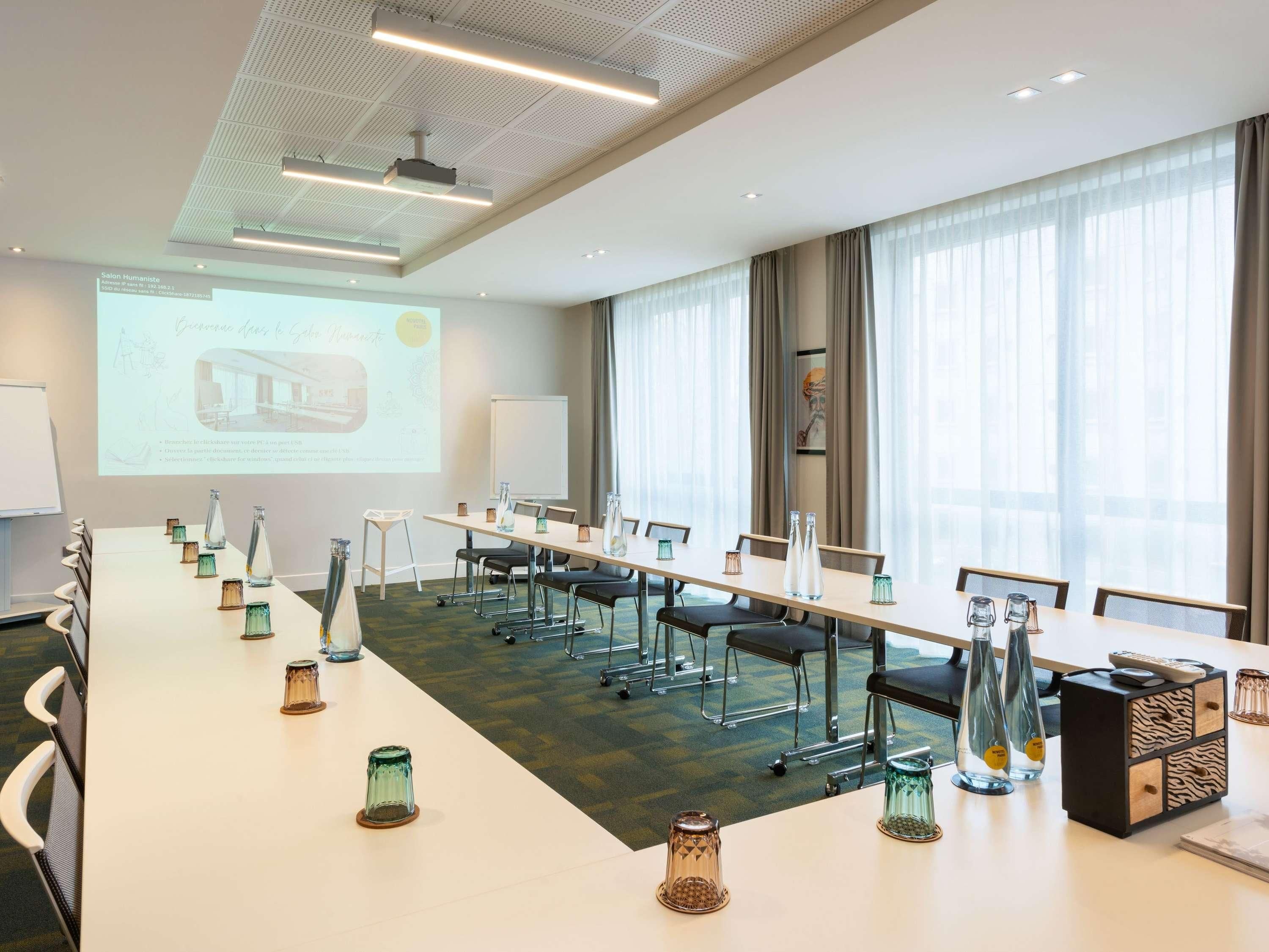 A conference room with a long table, chairs, a projector screen, and a large window with curtains.