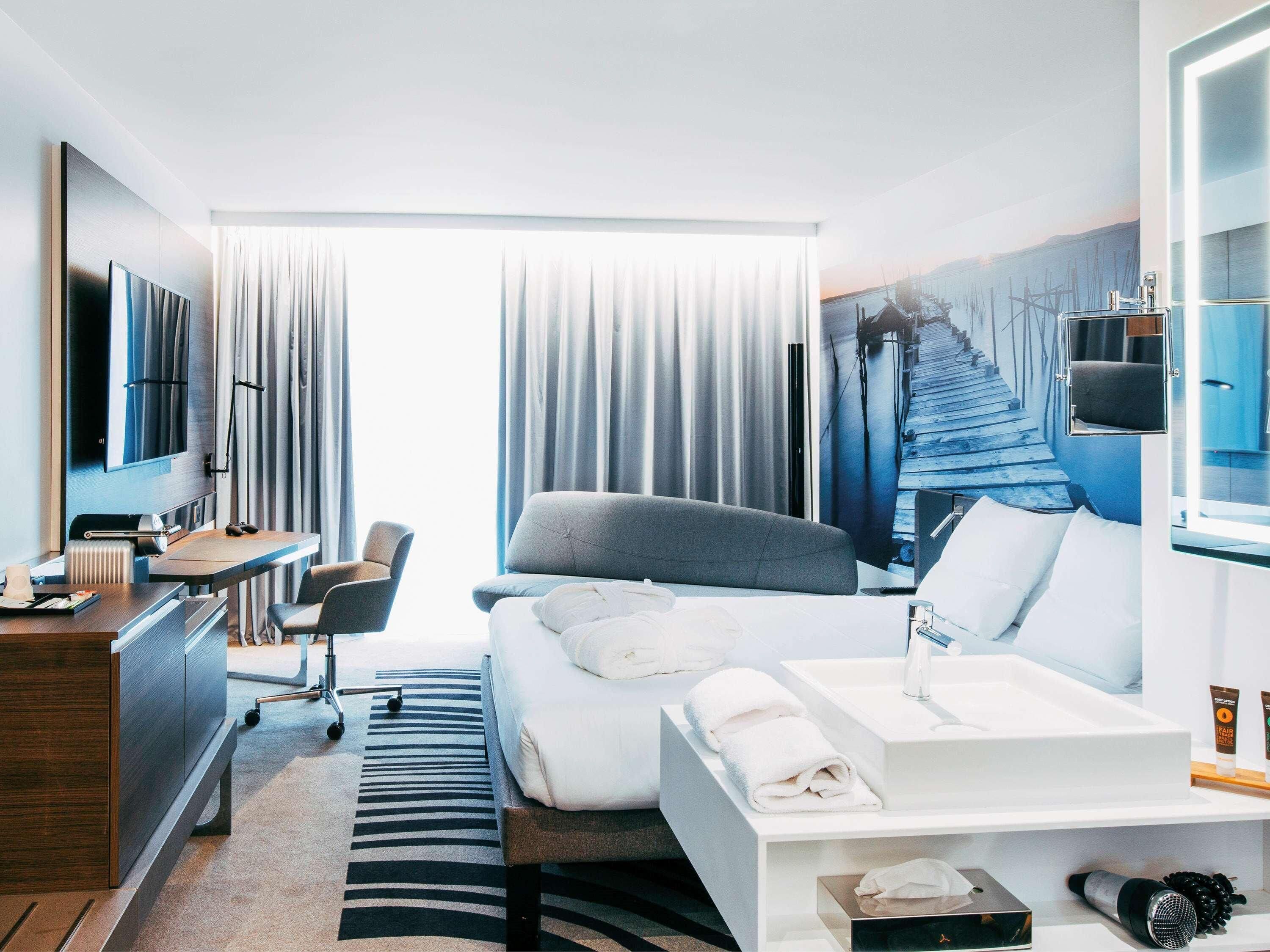 A hotel room with a bed, a desk, a chair, a TV, a painting, and a window with curtains.
