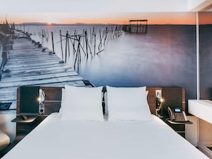 Habitació d'hotel amb un llit, taules de nit, un telèfon i vista al passeig marítim i al capvespre.