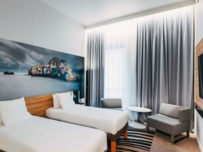 Habitación de hotel con dos camas, una mesita redonda pequeña, un sofá y un ventanal con cortinas.