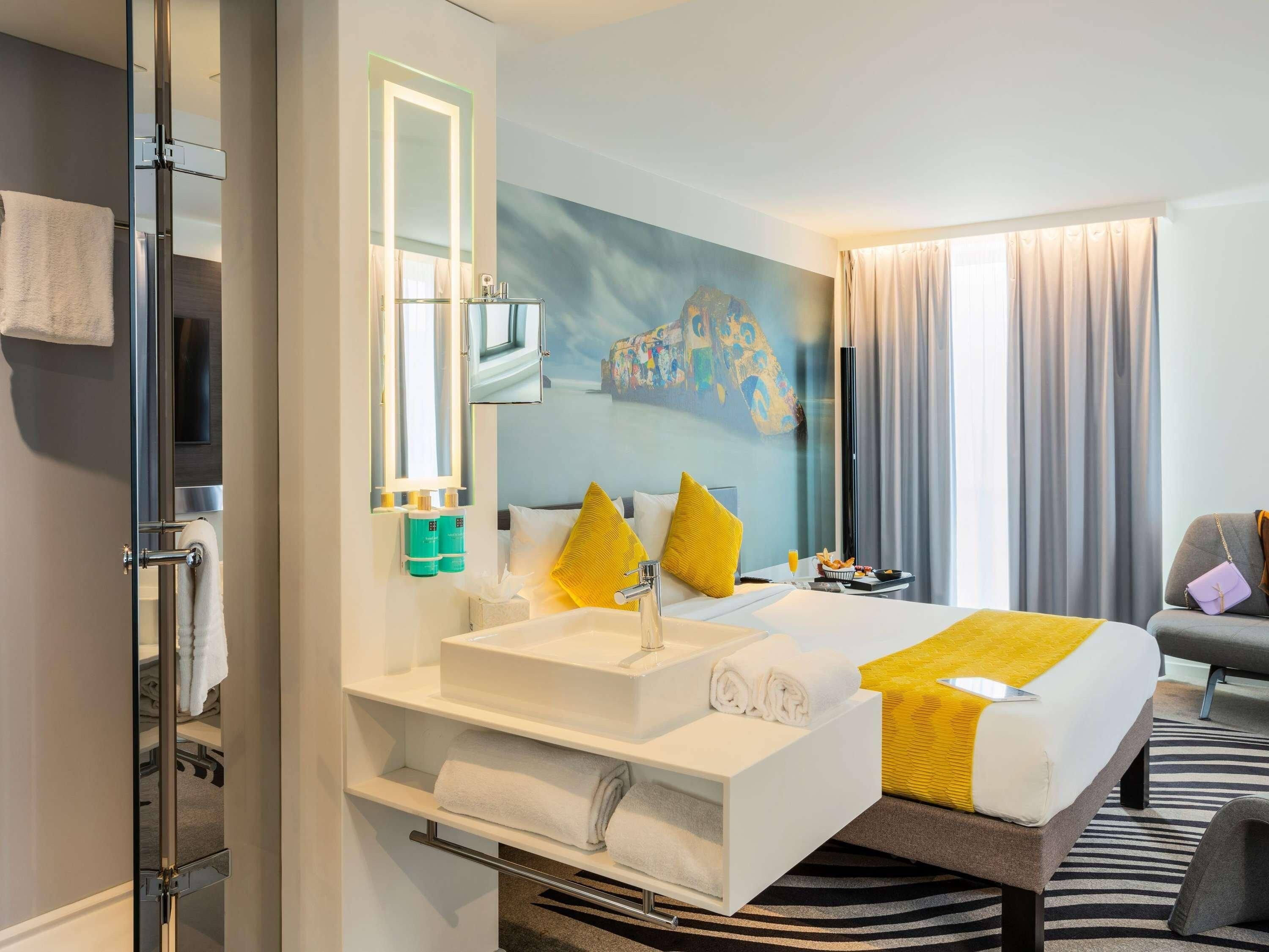A modern hotel room with a large bed, a white vanity with a mirror, a painting on the wall, and a view of the city.