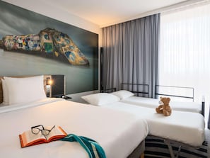 Una habitació d'hotel amb dos llits, un quadre abstracte gran a la paret, un os de peluix sobre un llit i un llibre amb ulleres sobre l'altre.