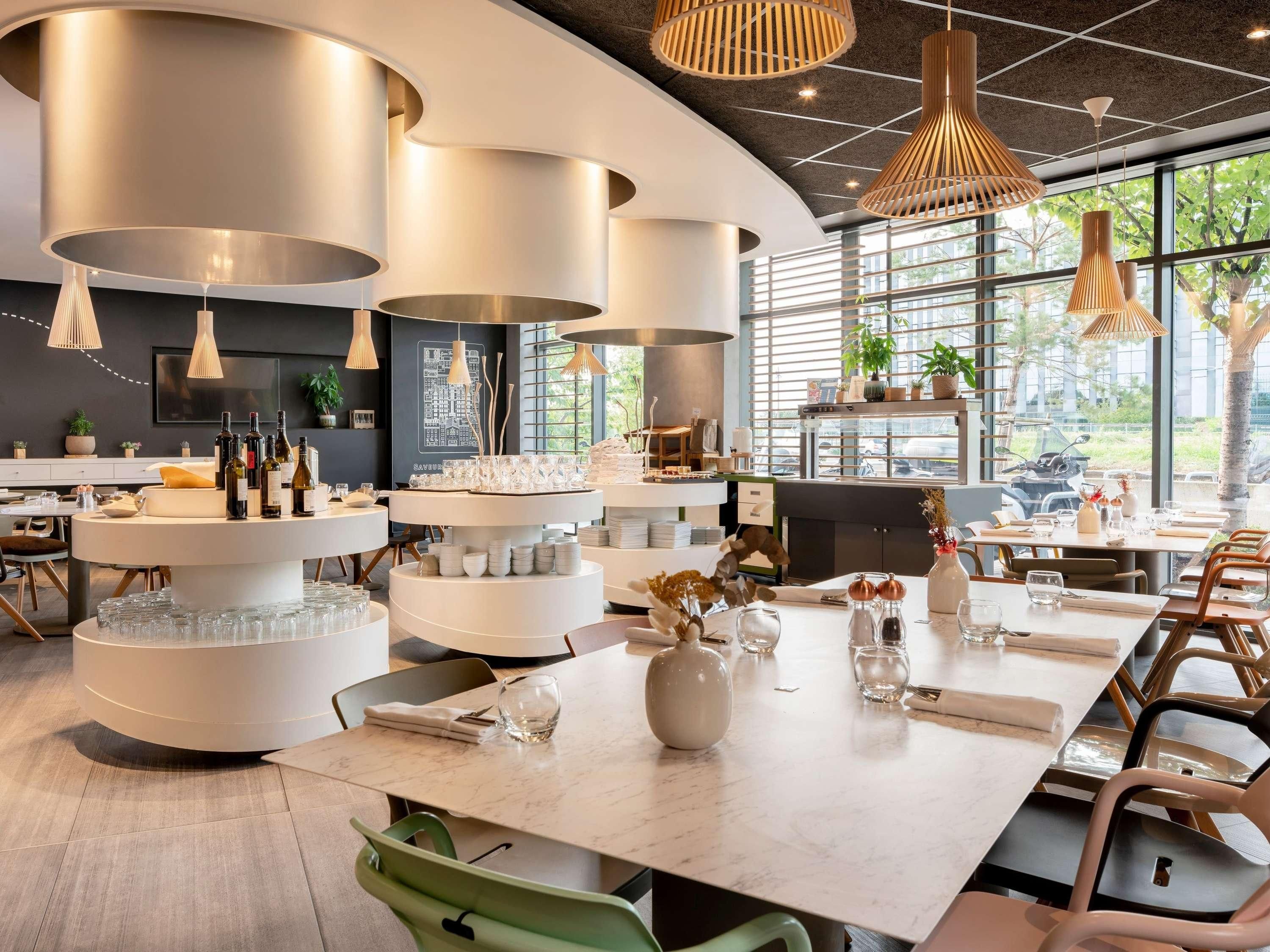 A modern dining area with round tables, hanging pendant lights, and a view of greenery outside.