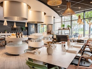 A modern dining area with round tables, hanging pendant lights, and a view of greenery outside. - Novotel Paris Coeur D'orly (Orly)