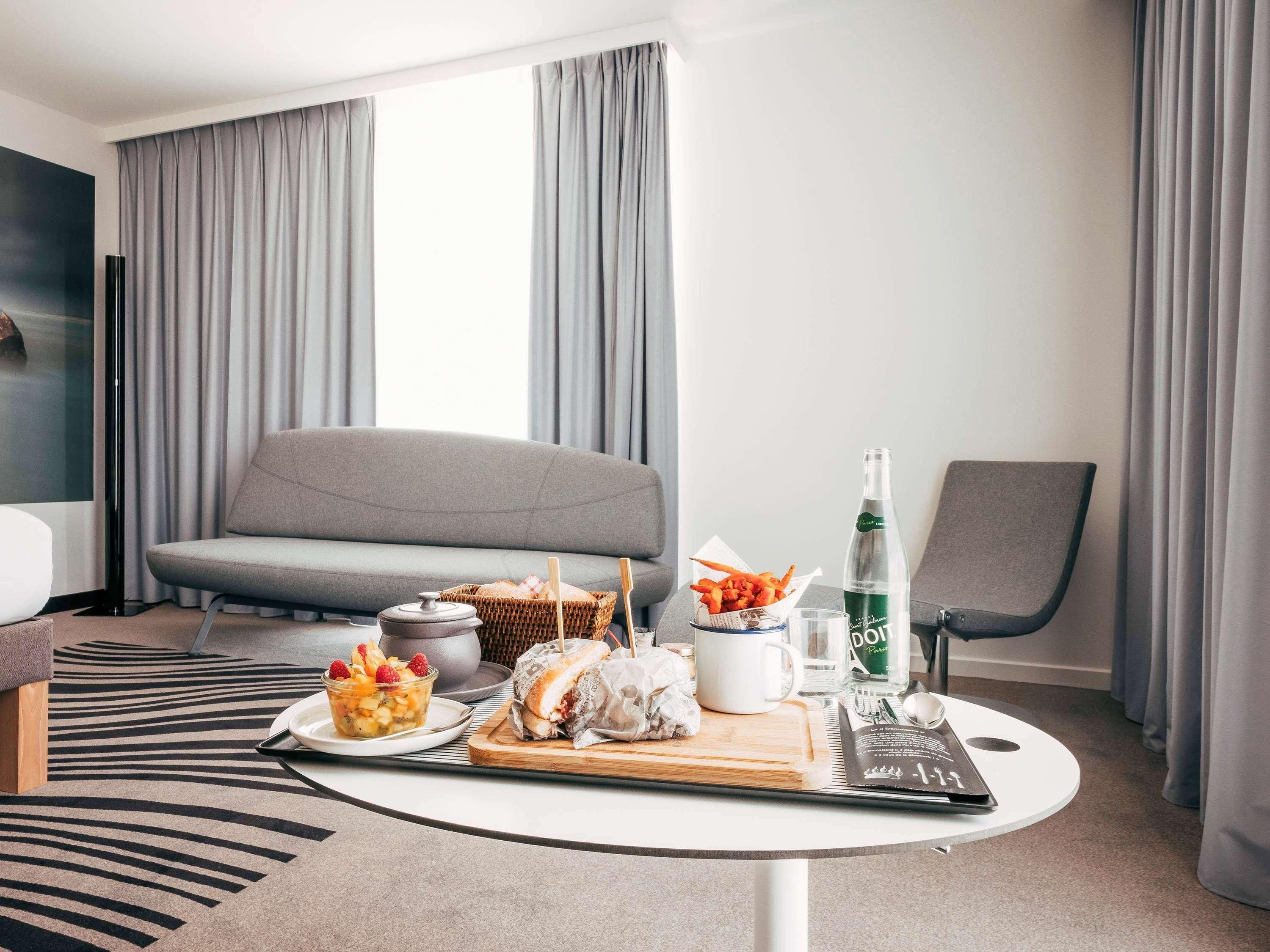 Een moderne hotelkamer met een grijze bank, een eettafel met eten en drinken, een stoel en een groot raam met gordijnen.