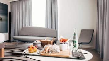 Een moderne hotelkamer met een grijze bank, een eettafel met eten en drinken, een stoel en een groot raam met gordijnen.